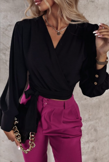 Frau in schwarzer Wickelbluse und pinker Hose mit goldener Kette vor grauem Hintergrund.