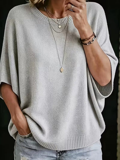Närbild framifrån på ljusgrå stickad topp med batwing-ärmar och ribbad halsringning, burna med lagda halsband och jeans. 1