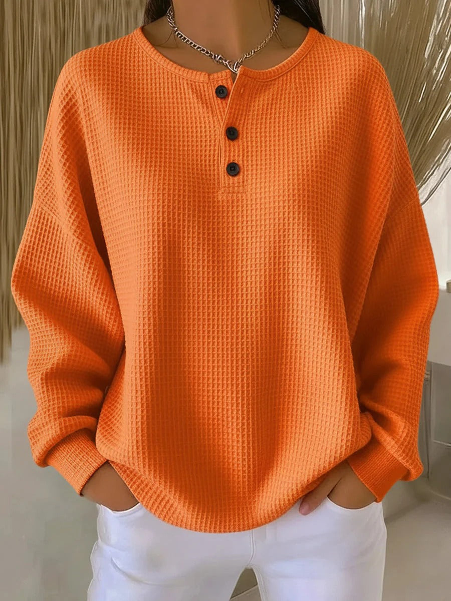 Enfärgad orange våffelstickad tröja med tre svarta knappar i halsringningen, buren av modell med vita byxor. 7