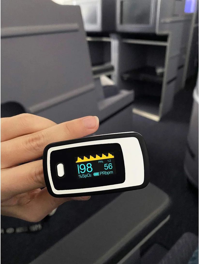 Person hält Finger-Pulsoximeter, zeigt Sauerstoffsättigung und Puls, im Flugzeug.