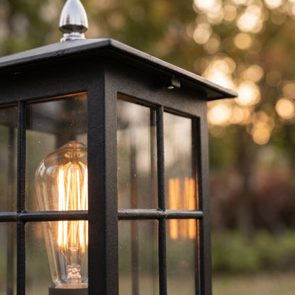 Närbild på lampans svarta, strukturerade ram, rutindelade glas och lysande glödlampa med glödtråd, med skimrande bokeh i bakgrunden. 5
