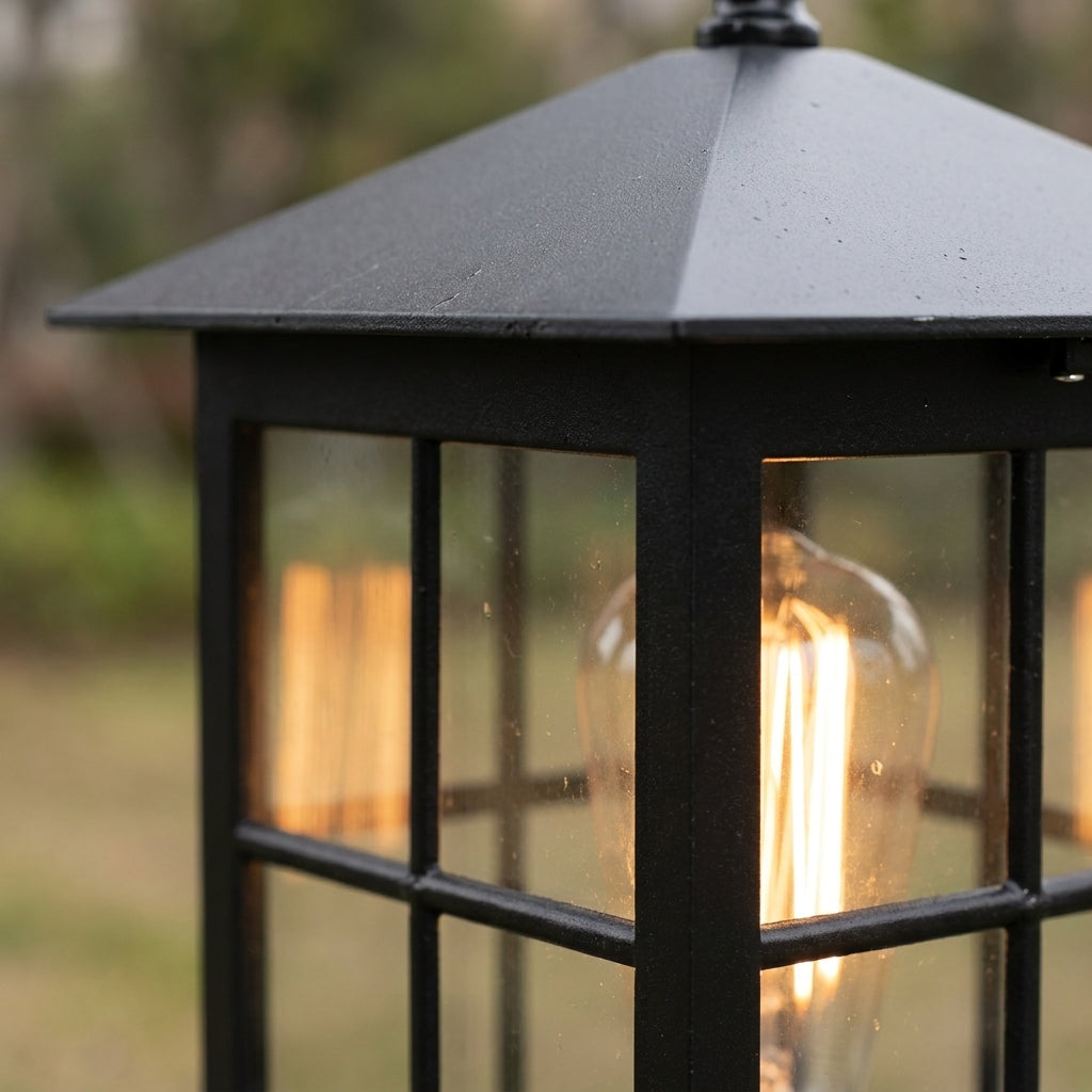 Närbild på lampans sluttande svarta tak och rutindelade glas med en lysande glödlampa bakom, utomhus med suddig bakgrund. 6