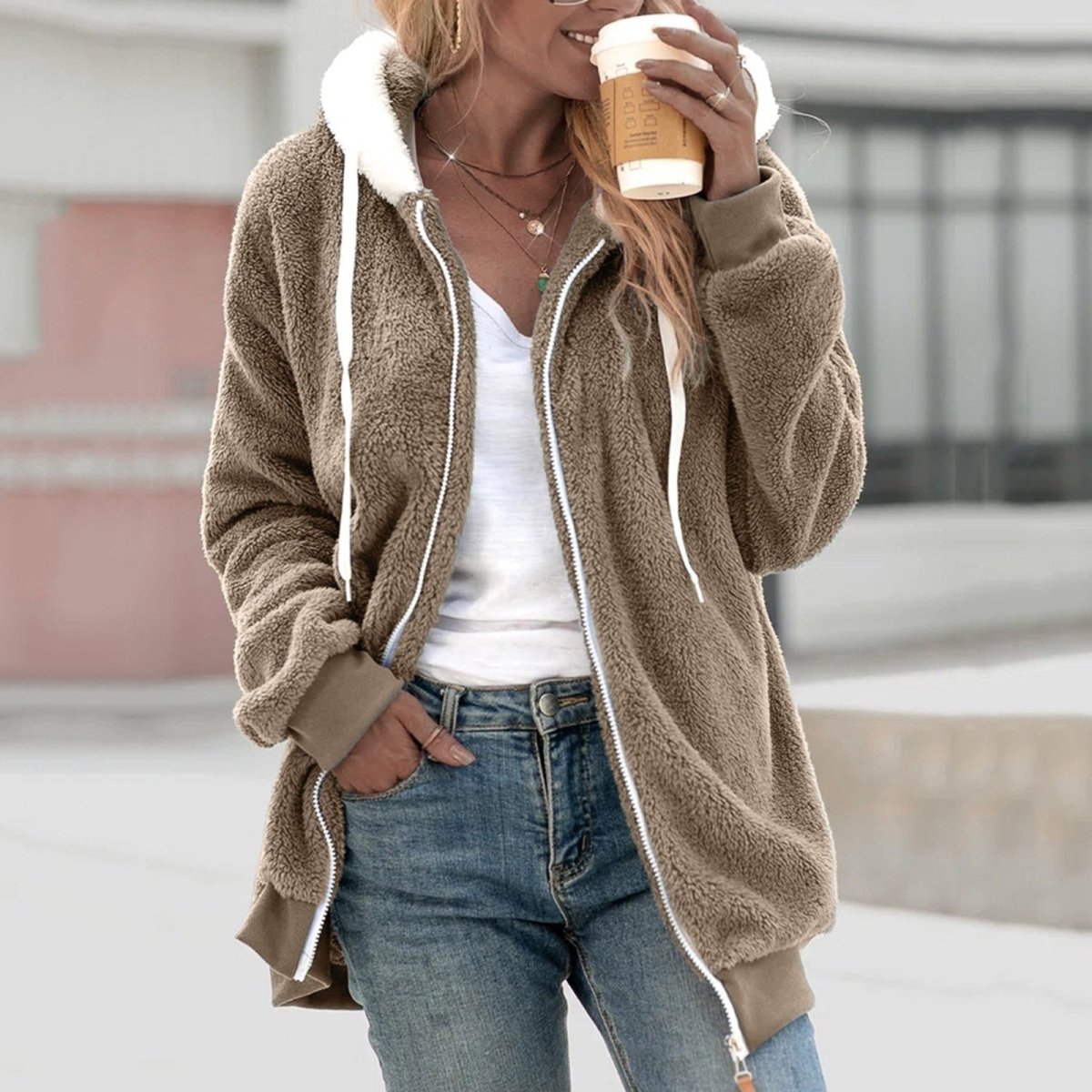 Frau in brauner Teddyweste mit Reißverschluss, Kapuze, hält Kaffeebecher, lässige Mode.