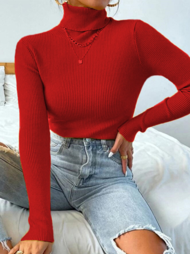Frau in rotem Rollkragenpullover, gerippt, kombiniert mit blauen Jeans, sitzt auf einem Bett.