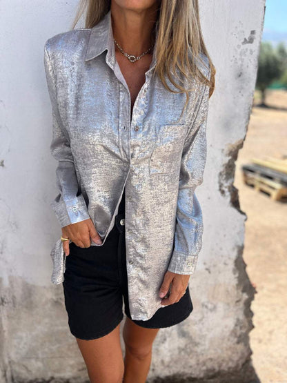 Frau in silberner Bluse und schwarzen Shorts vor rustikalem Hintergrund, modisch, elegant.