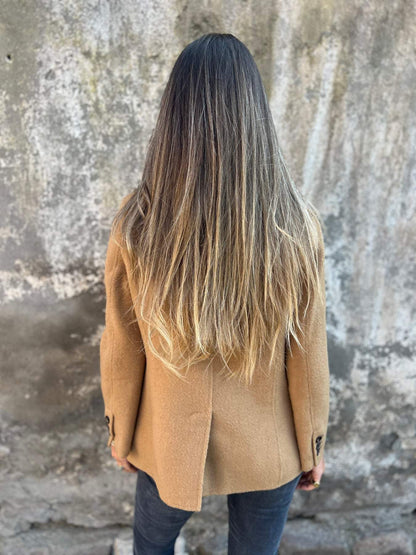 Frau mit langen, glatten Haaren in braunem Mantel vor Betonwand.