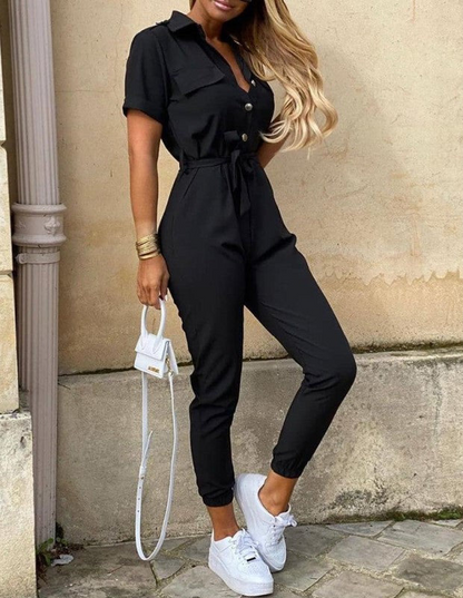 Frau in schwarzem Jumpsuit mit Gürtel, kurzen Ärmeln, weißen Sneakers und kleiner Handtasche.
