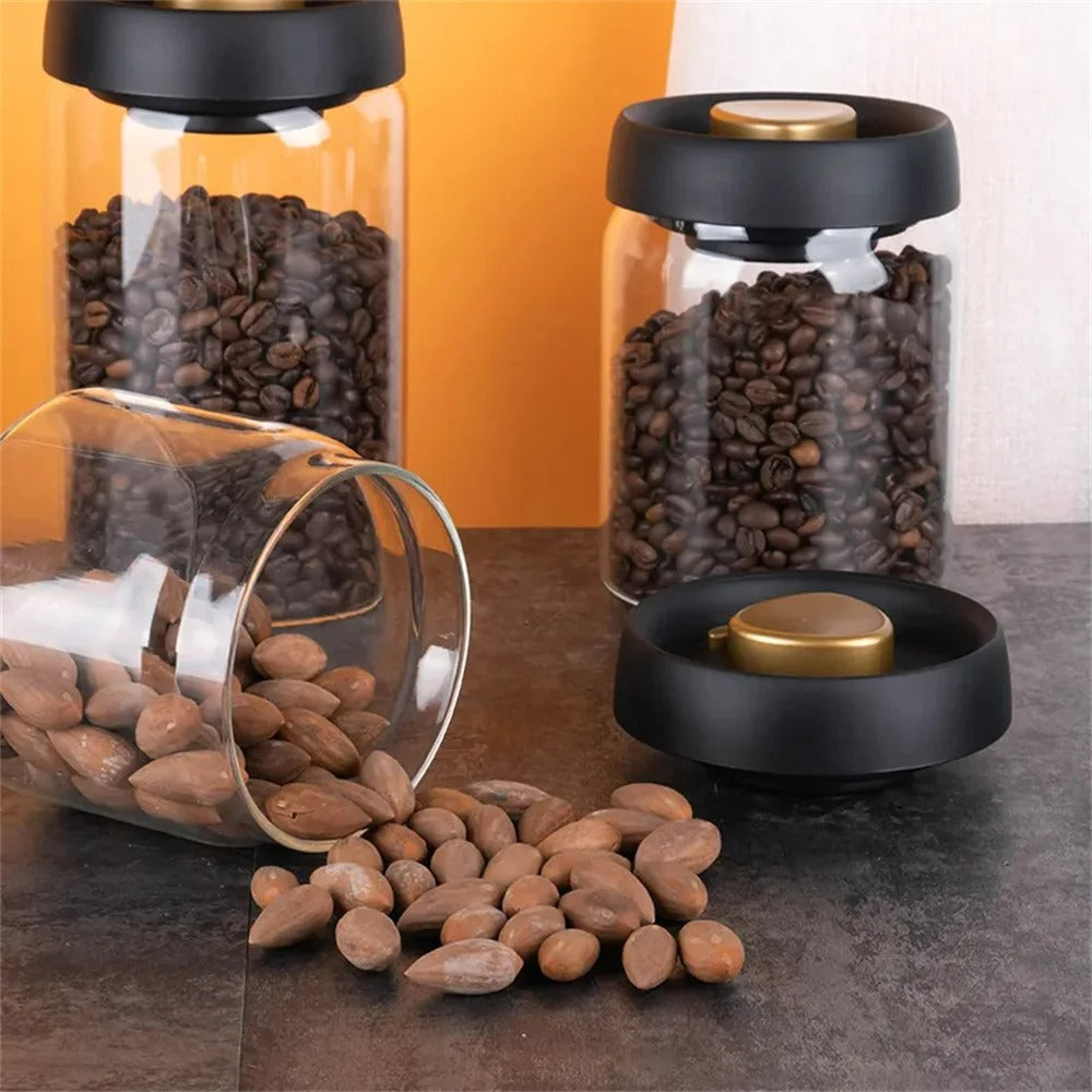 Glasbehälter mit schwarzem Deckel, gefüllt mit Kaffeebohnen, auf grauem Tisch vor orangefarbener Wand.