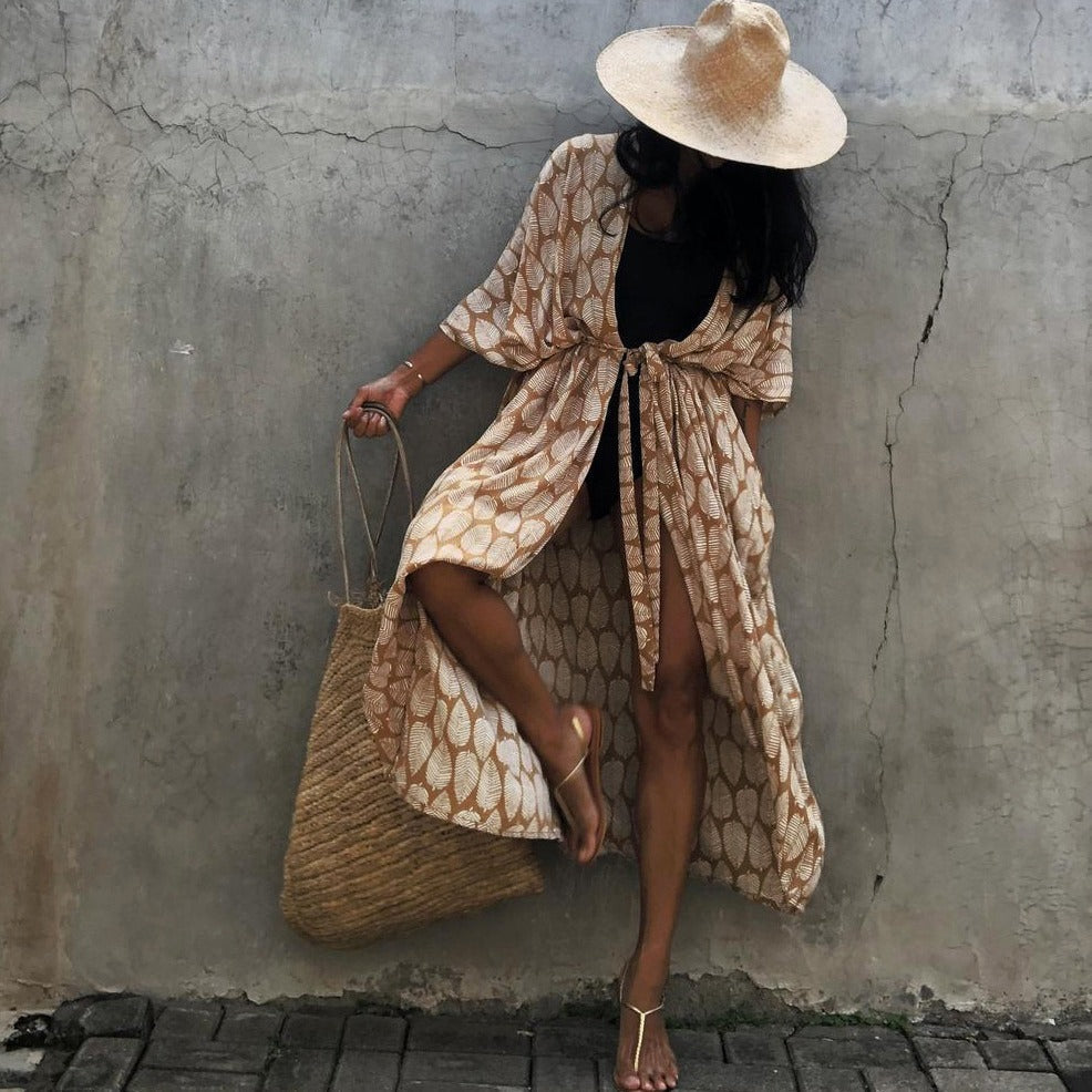 Frau in langem, gemustertem Kimono mit Strohhut und Tasche vor Betonwand.