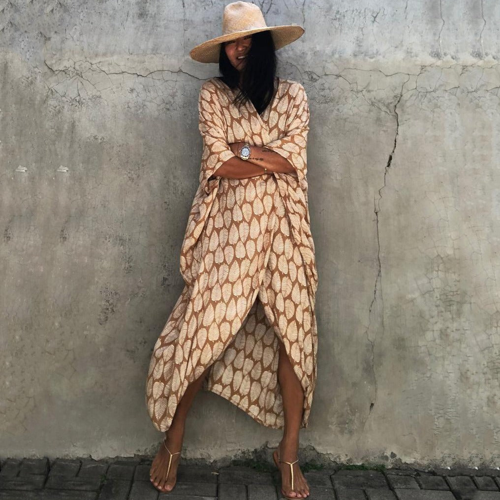 Frau in beige-gemustertem Kaftan und Strohhut vor Betonwand, Sommermode, lässiger Stil.