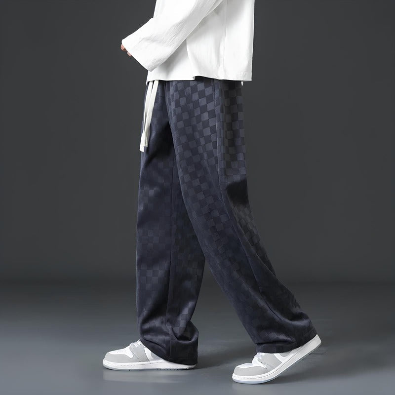 Mann in karierten, schwarzen Hosen und weißen Sneakers vor grauem Hintergrund.