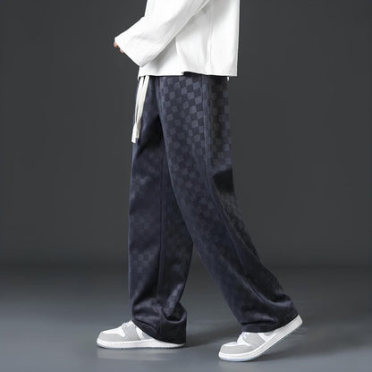 Mann in karierten, schwarzen Hosen und weißen Sneakers vor grauem Hintergrund.