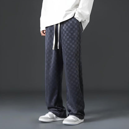Mann in karierten, dunkelgrauen Jogginghosen und weißen Sneakers vor grauem Hintergrund.