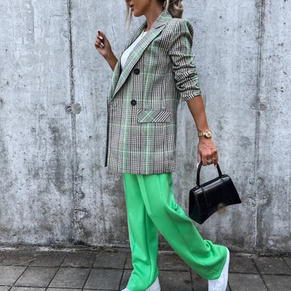 Frau in kariertem Blazer, grüner Hose, weißem Shirt, mit schwarzer Handtasche vor Betonwand.