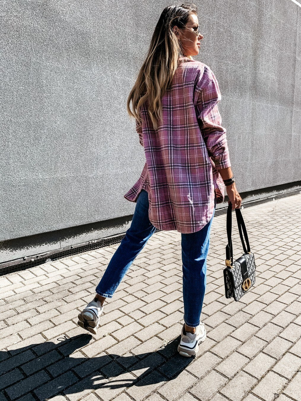 Frau in rosa kariertem Hemd, Jeans und Sneakers, trägt schwarze Handtasche, geht auf Bürgersteig.