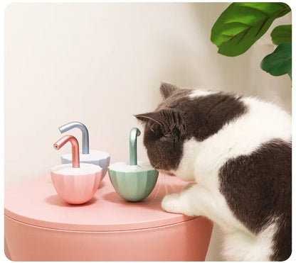 Katze neben bunten, modernen Trinkbrunnen für Haustiere auf rosa Tisch, Pflanzen im Hintergrund.