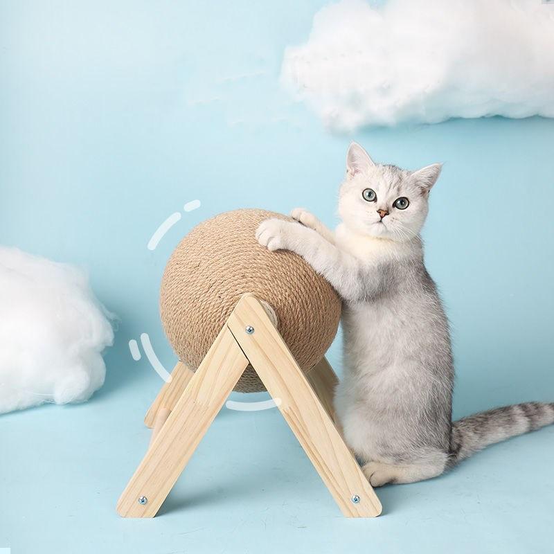 Graue Katze mit Kratzball aus Sisal auf Holzgestell vor blauem Hintergrund mit Wolken.