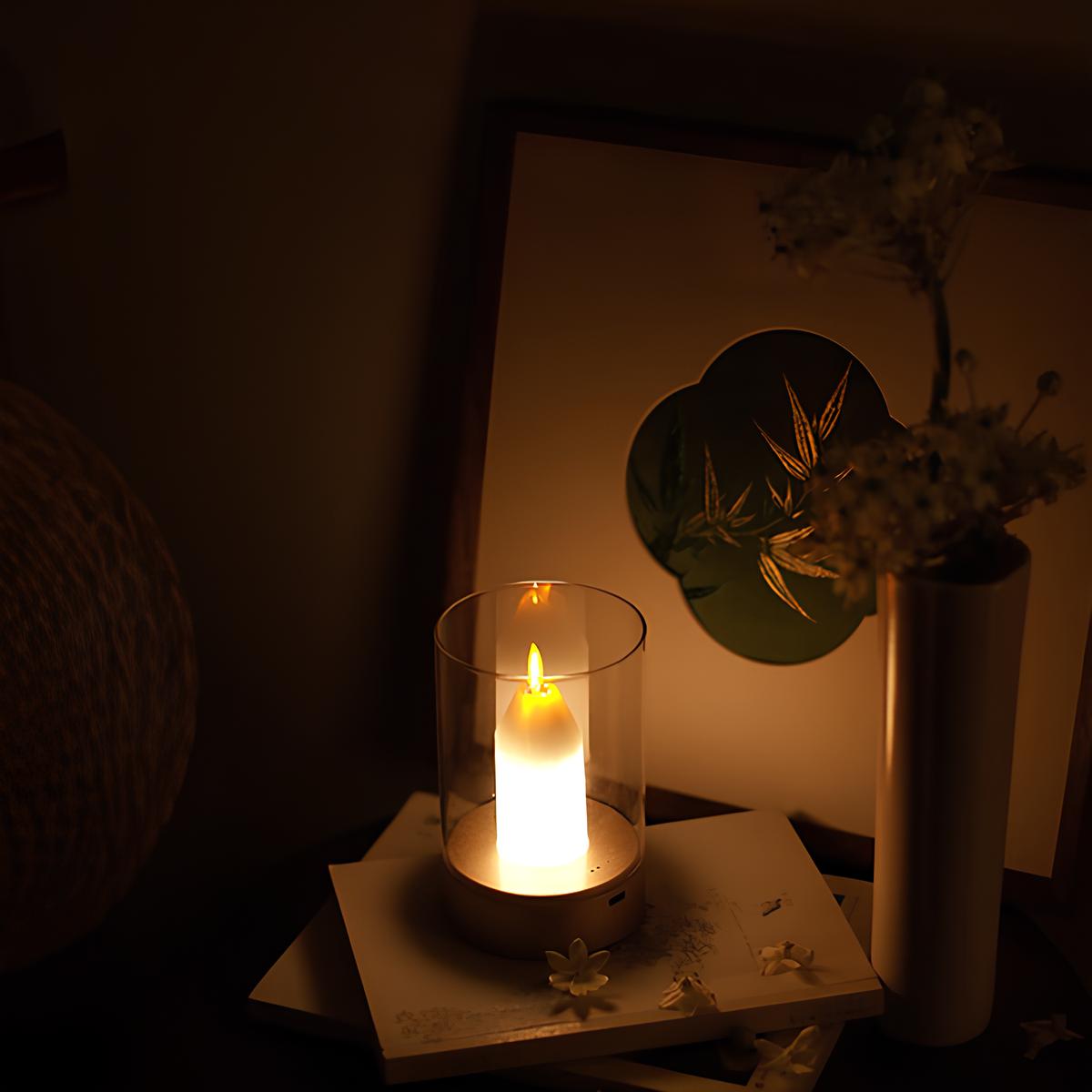 Beleuchtete Kerze im Glas auf Tisch, umgeben von Blumen und Kunstwerk, gemütliche Atmosphäre.