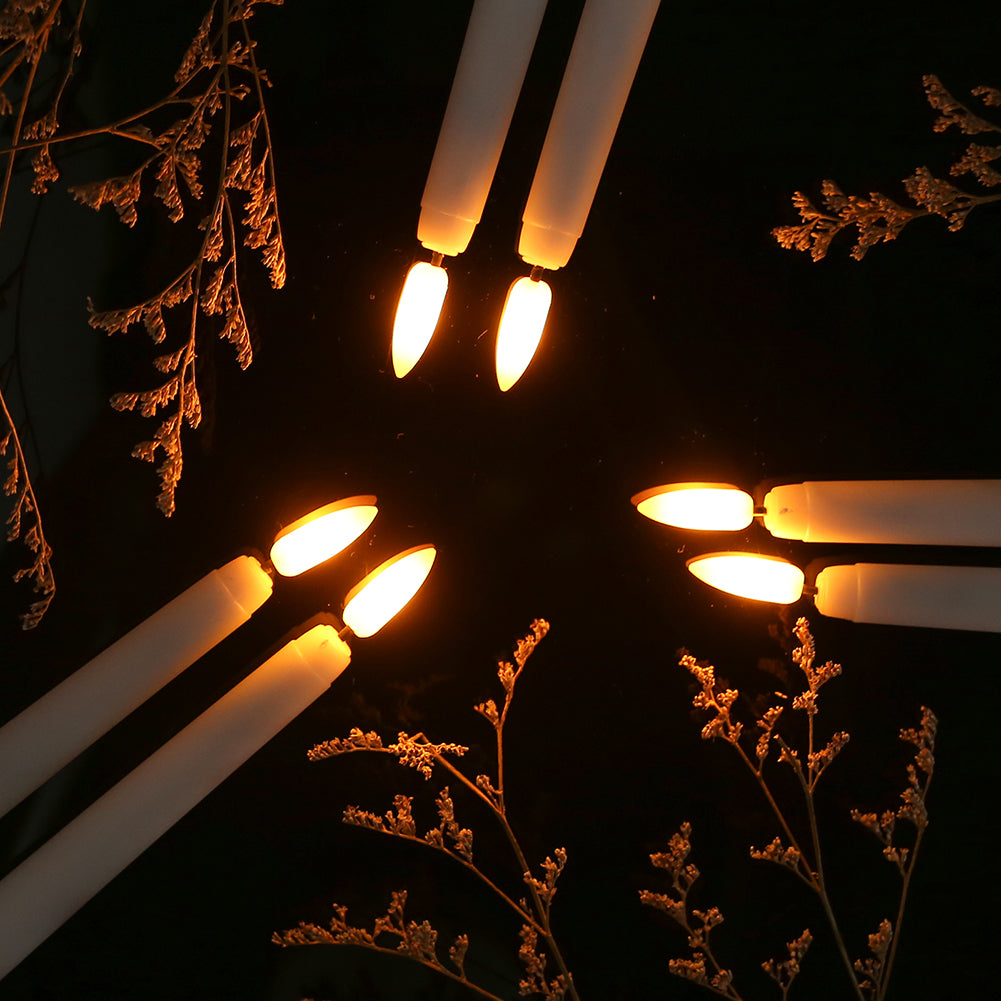 Sechs LED-Kerzen, warmweiß leuchtend, umgeben von getrockneten Zweigen auf schwarzem Hintergrund.