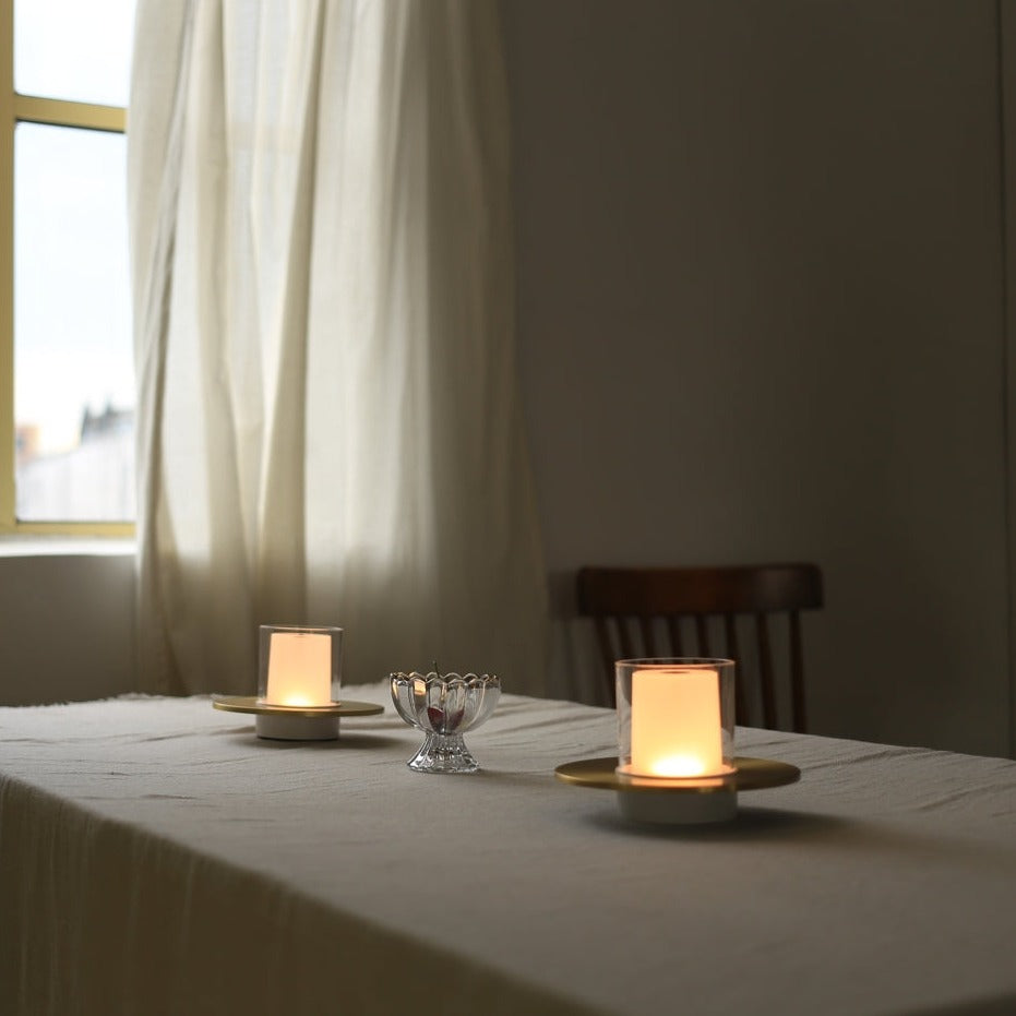Zwei brennende Kerzen auf Tisch mit weißer Tischdecke, Fenster im Hintergrund, gemütliche Atmosphäre.