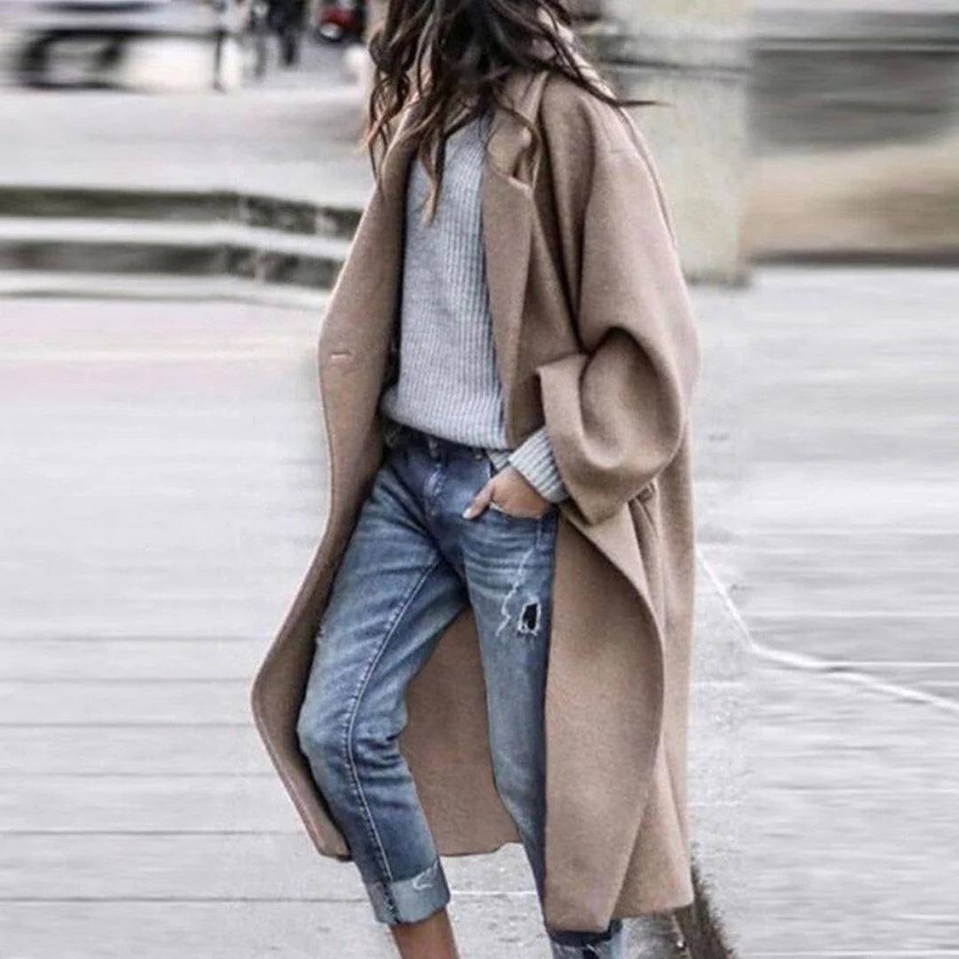 Frau in khakifarbenem Mantel, grauem Pullover und Jeans auf der Straße.