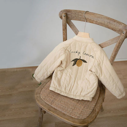 Beige Kinderjacke mit "Lucky Lemon"-Stickerei, hängend auf einem Holzstuhl.