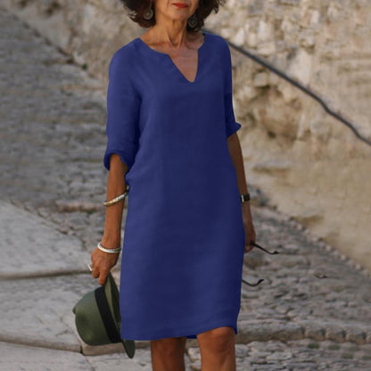 Frau in blauem Sommerkleid, V-Ausschnitt, auf Kopfsteinpflasterweg, hält Hut in der Hand.