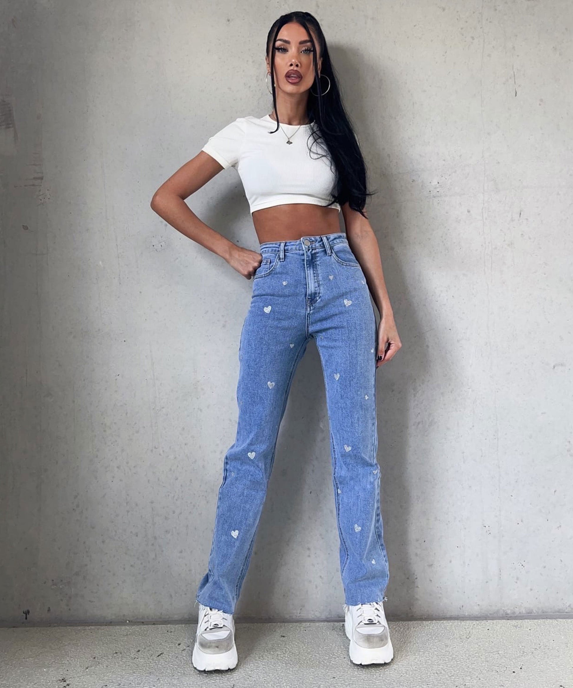Frau in weißem Crop-Top und blauer Jeans mit Herzmuster, weiße Sneaker, grauer Hintergrund.