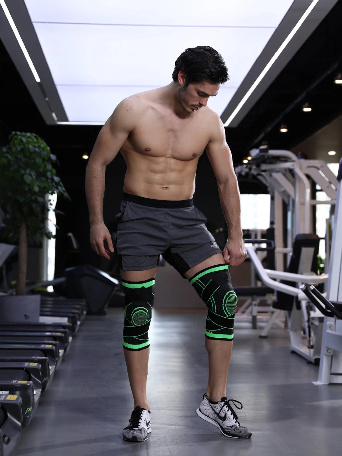 Mann im Fitnessstudio mit grauen Shorts und grünen Kniebandagen, trainiert auf Laufband.