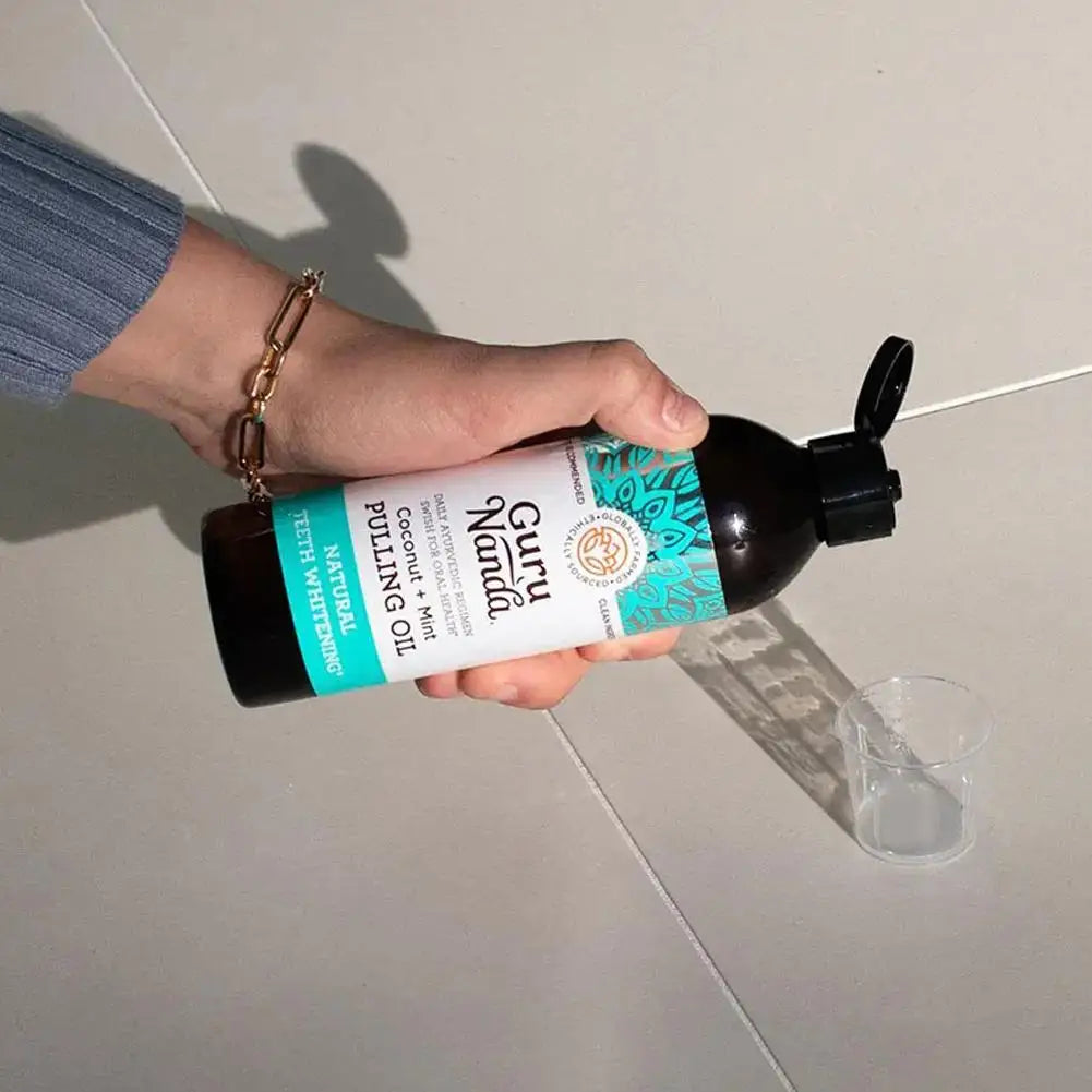 Hand hält GuruNanda Ölziehflasche, Kokosnuss-Minz-Geschmack, 473 ml, auf Fliesenhintergrund.