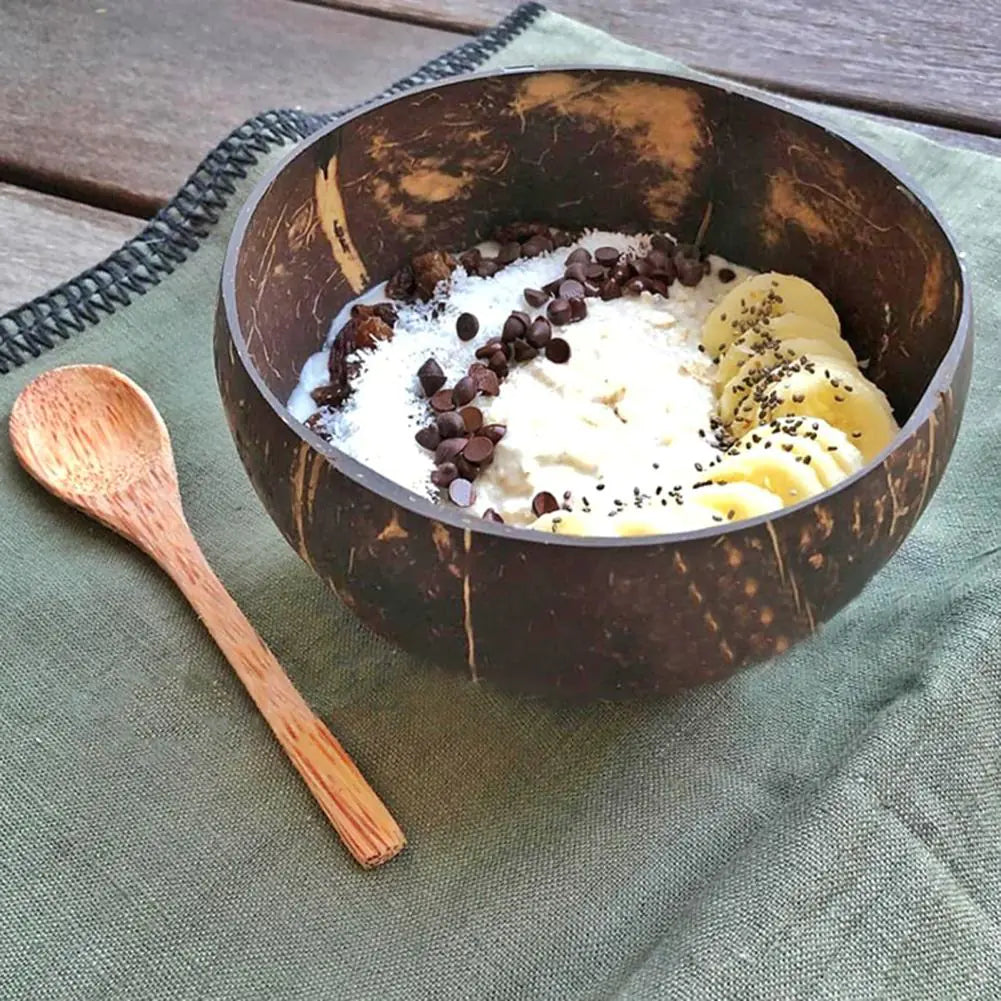 Kokosnussschale mit Joghurt, Bananen, Schokostückchen auf grünem Tuch, Holzlöffel daneben.