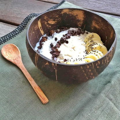 Kokosnussschale mit Joghurt, Bananen, Schokostückchen auf grünem Tuch, Holzlöffel daneben.