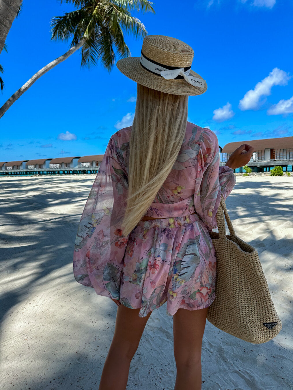 Frau in floralem Sommerkleid und Strohhut am Strand, trägt geflochtene Tasche, Palmen im Hintergrund.