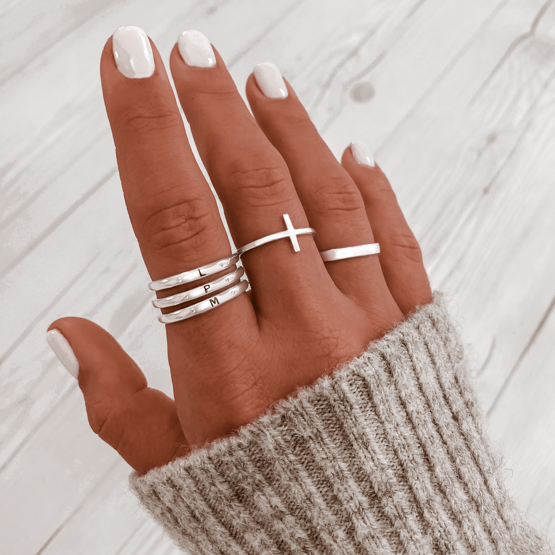 Hand mit silbernen Ringen, Kreuzring, Buchstabenringe, grauer Strickpullover, Nagellack.