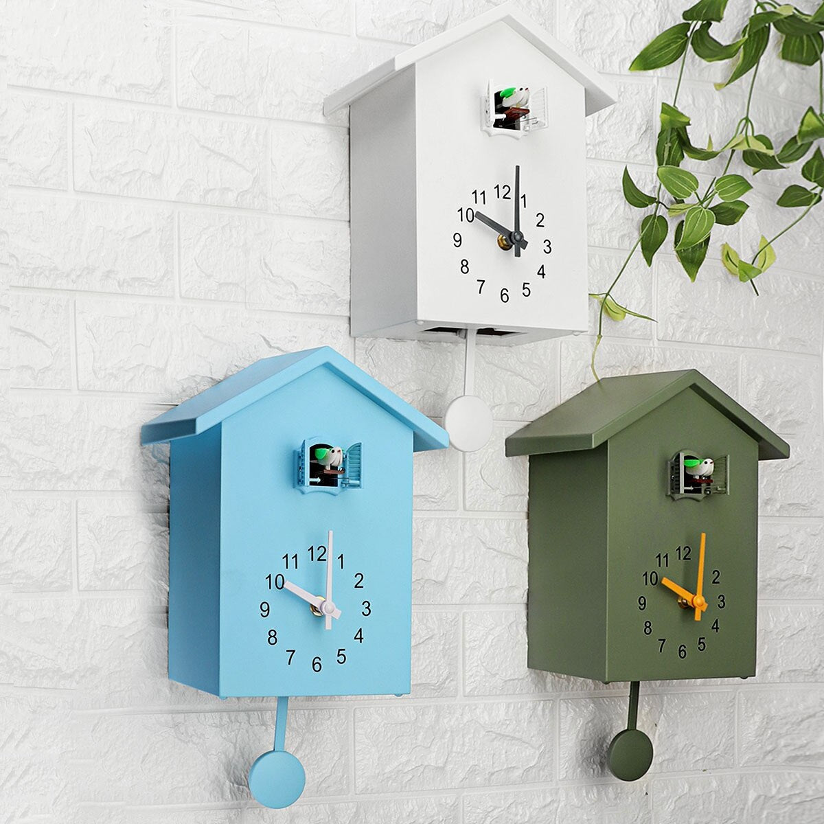 Drei Kuckucksuhren in Blau, Weiß und Grün an Ziegelwand, modernes Design, hängende Pflanzen.