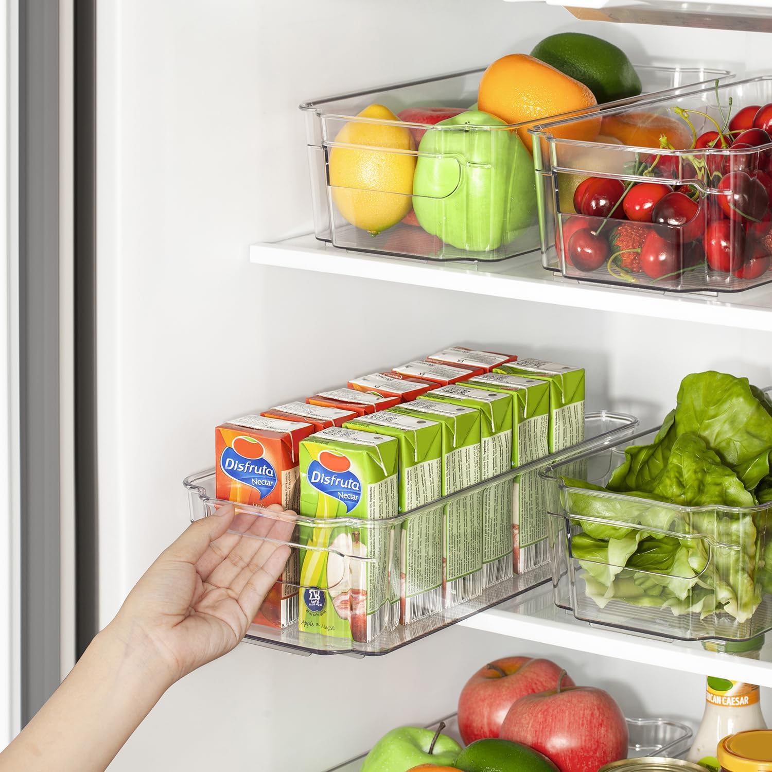 Hand greift nach Saftpackung im transparenten Kühlschrank-Organizer mit Obst und Gemüse.