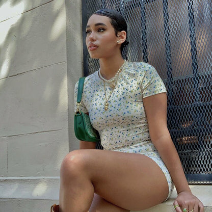 Frau in geblümtem Kleid sitzt draußen, trägt grüne Tasche, Sommermode, urbaner Hintergrund.