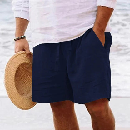 Mann in weißem Hemd und blauer Shorts am Strand, hält Strohhut in der Hand.