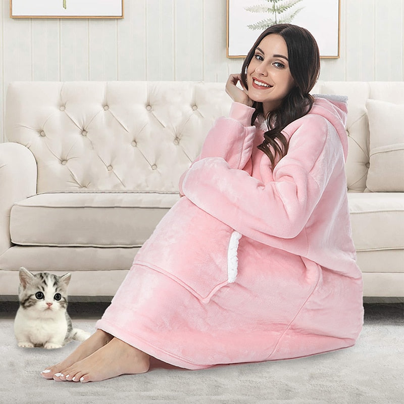 Frau in rosa Kuscheldecke mit Kapuze sitzt auf Sofa, Katze daneben, gemütliches Wohnzimmer.