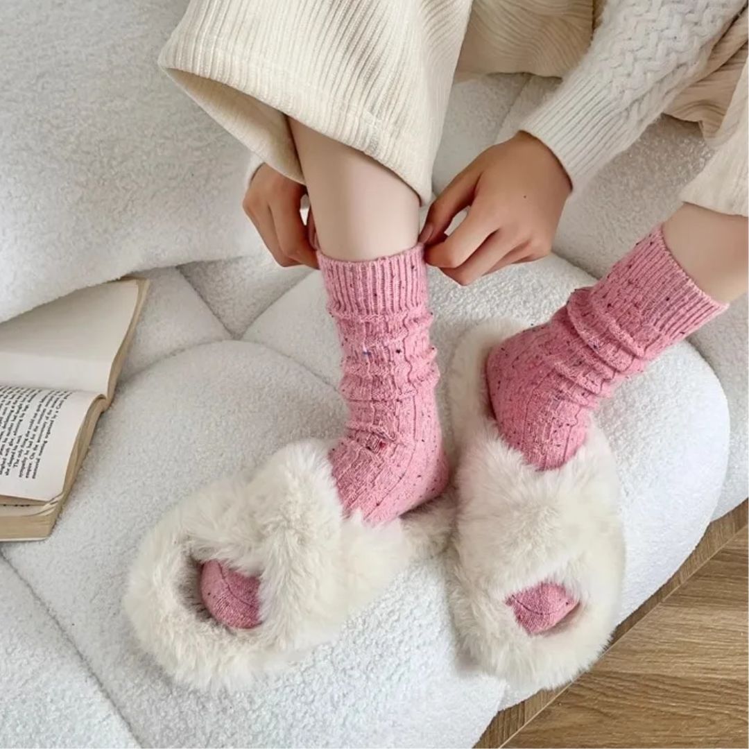 Frau mit rosa Socken und weißen Plüschpantoffeln auf einem weißen Sofa, gemütliche Wintermode.
