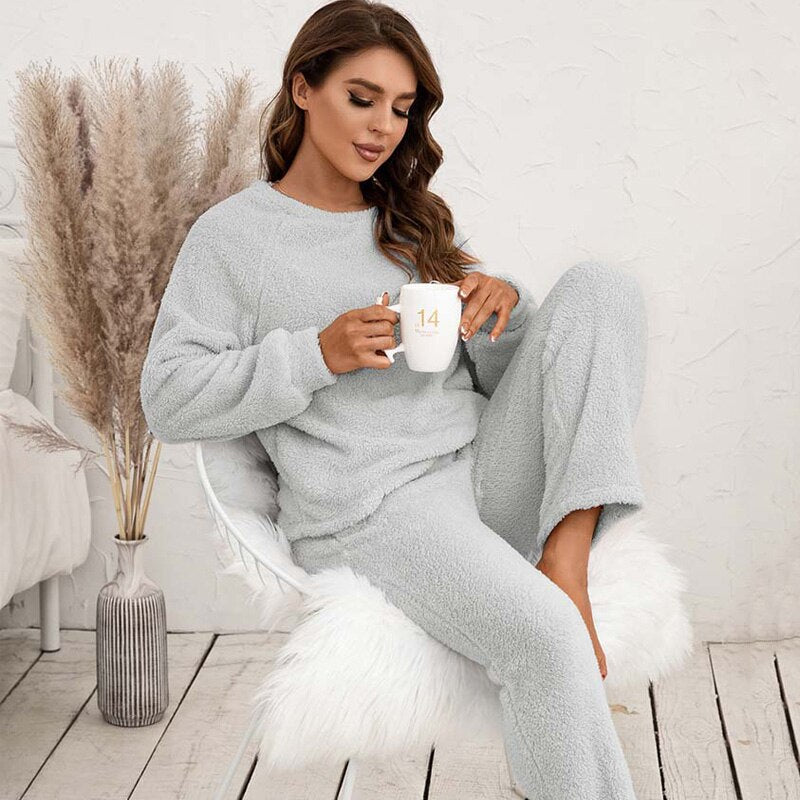 Frau in grauem Fleece-Pyjama mit Tasse, sitzt auf weißem Stuhl, gemütliche Wohnzimmeratmosphäre.