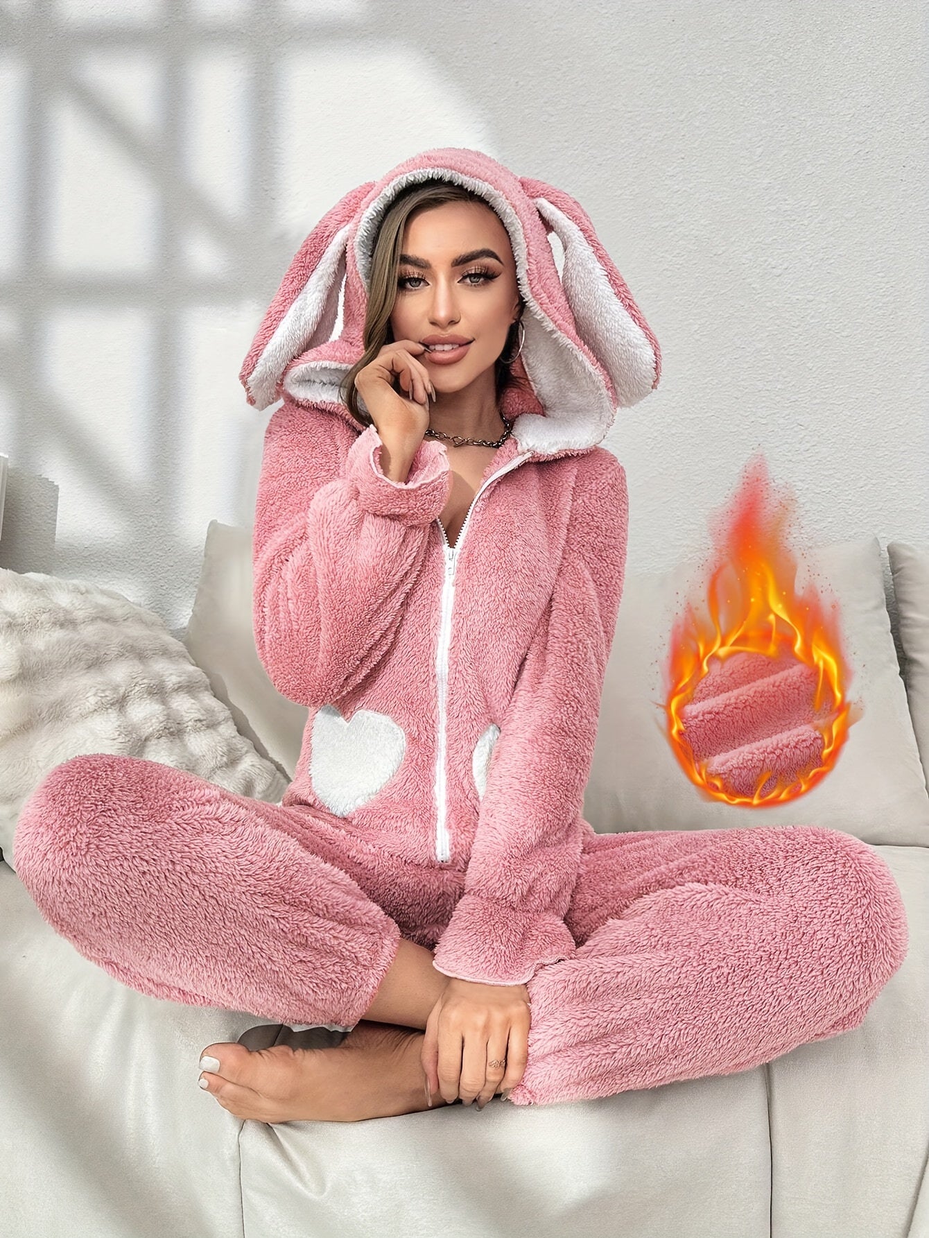 Frau in rosa Plüsch-Hasen-Einteiler mit Kapuze, Herzmuster, gemütlich, auf Sofa sitzend.