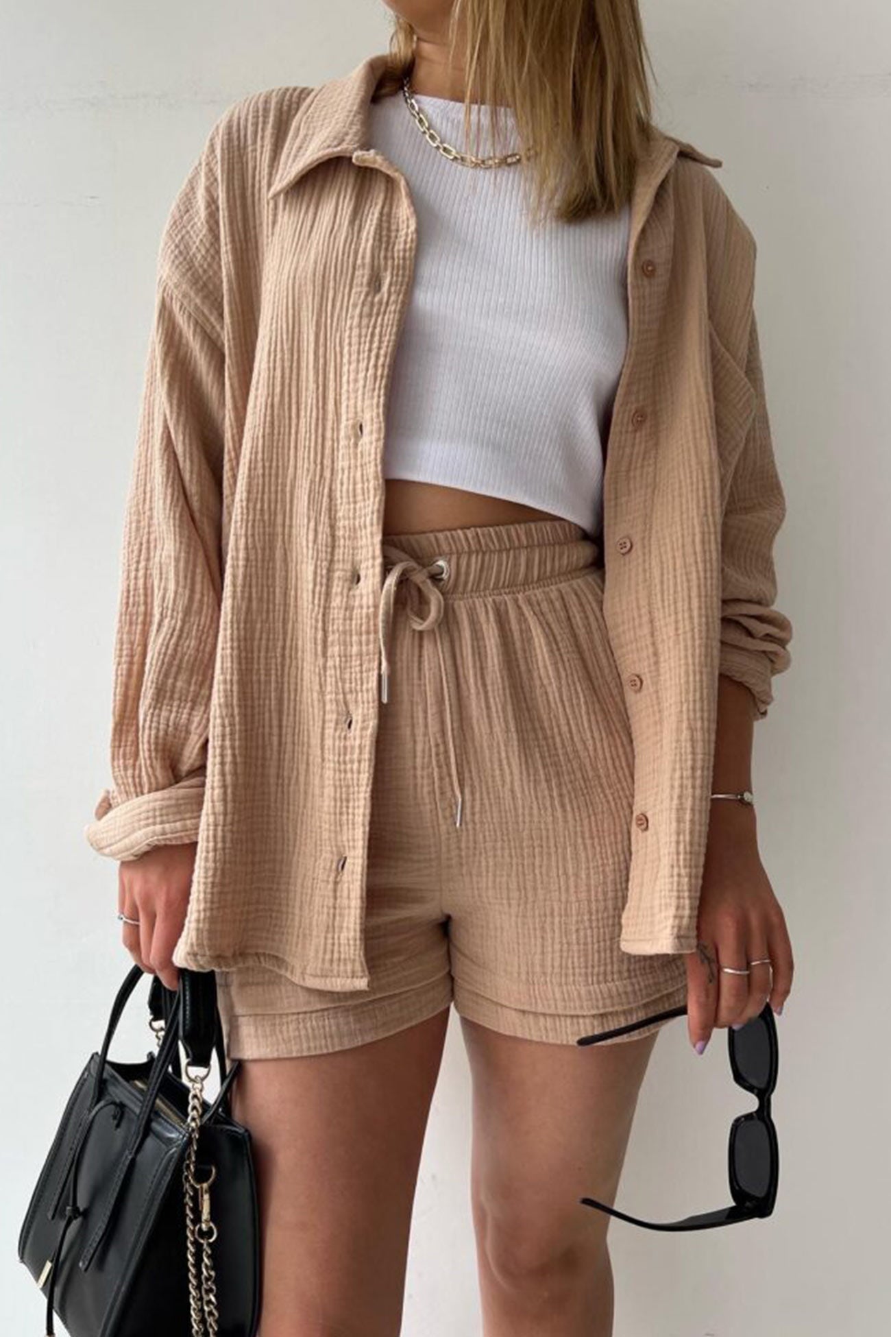 Frau in beigem Langarmhemd und Shorts, weißes Top, schwarze Tasche, Sonnenbrille in der Hand.