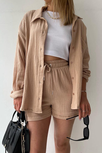 Frau in beigem Langarmhemd und Shorts, weißes Top, schwarze Tasche, Sonnenbrille in der Hand.