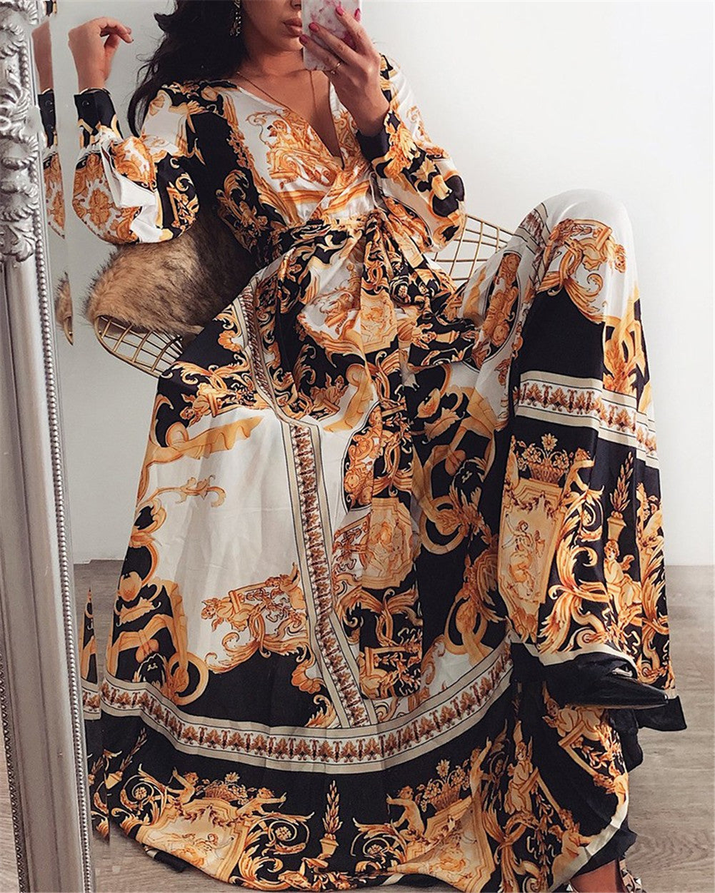 Frau in langem, gemustertem Maxikleid mit barockem Design, Schwarz-Gold-Weiß, sitzt auf Stuhl.