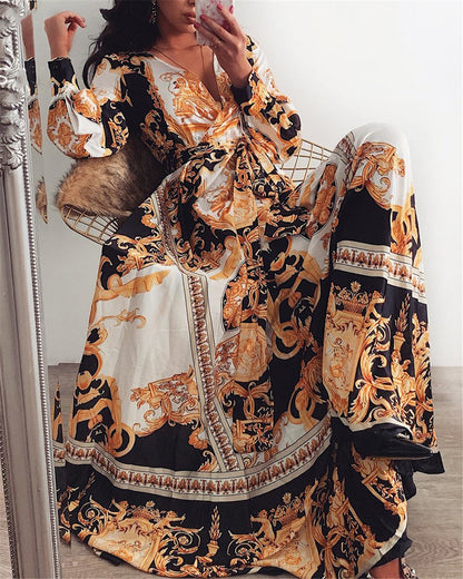 Frau in langem, gemustertem Maxikleid mit barockem Design, Schwarz-Gold-Weiß, sitzt auf Stuhl.