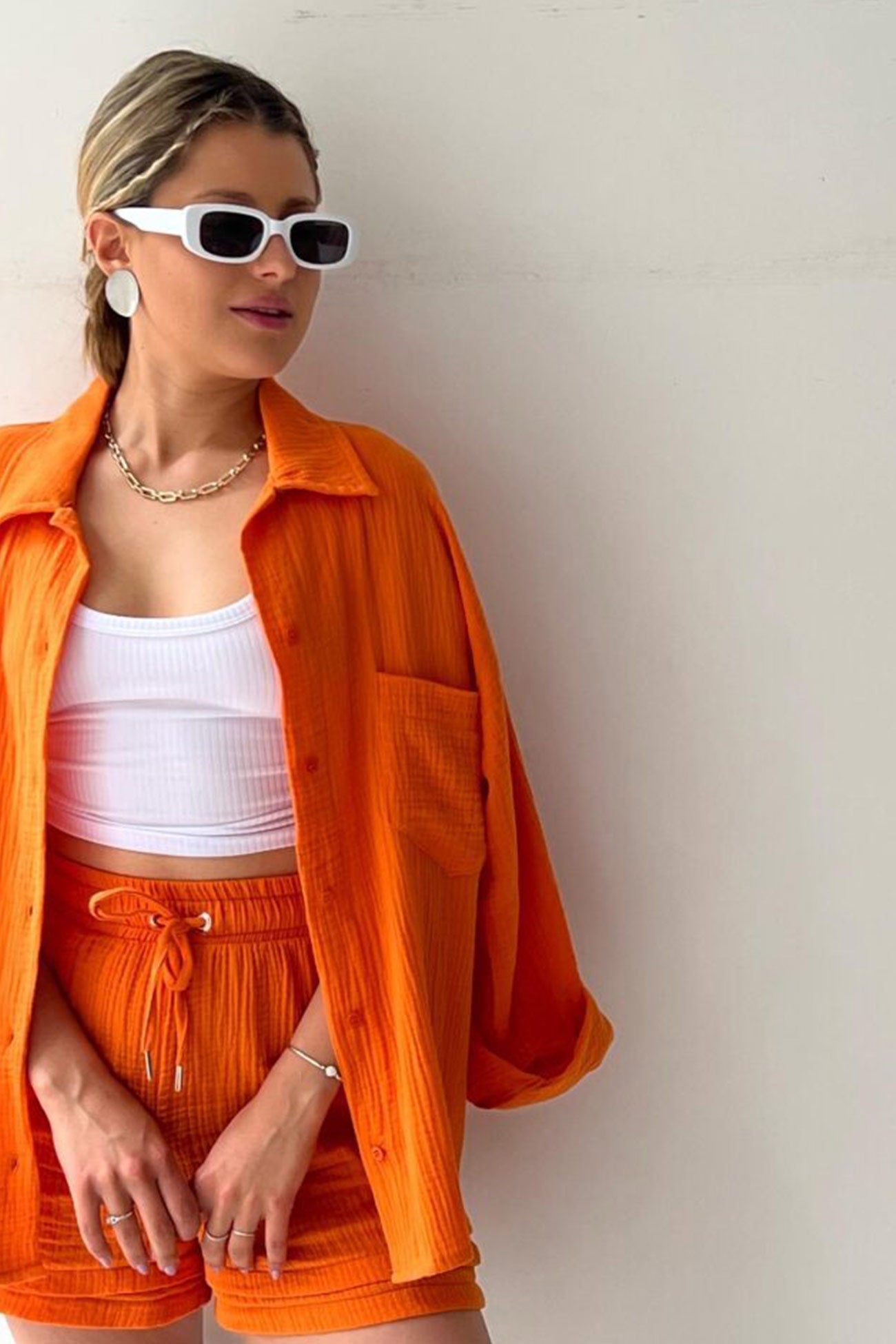 Frau in orangefarbenem Langarmhemd und Shorts, weiße Sonnenbrille, modisches Sommeroutfit.