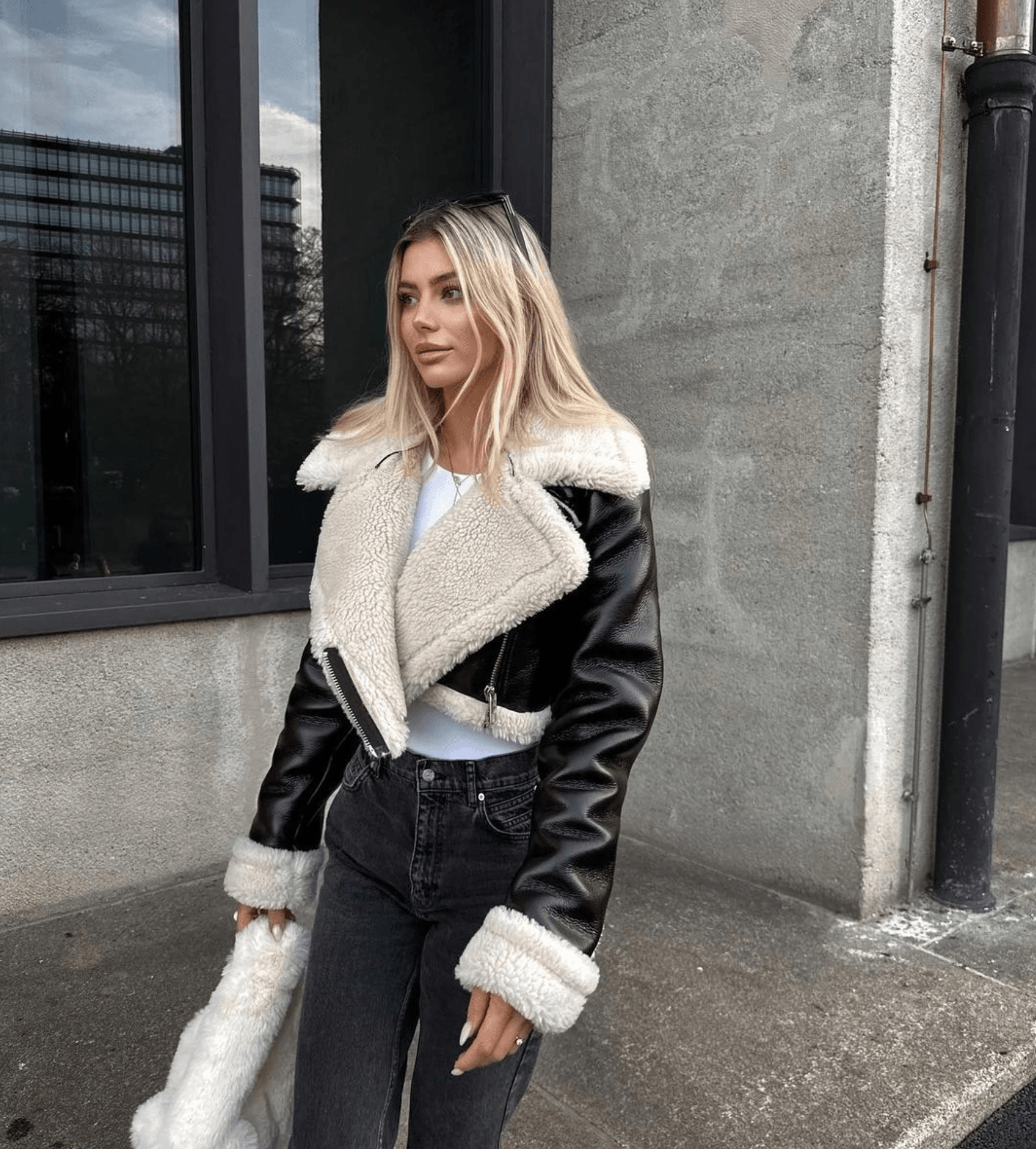 Frau in schwarzer Leder-Teddyjacke, weißem Top und Jeans vor Gebäude.