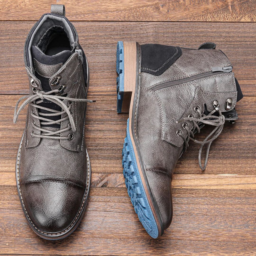 Graue Herrenstiefel aus Leder mit blauer Sohle, Schnürung und Reißverschluss auf Holzboden.