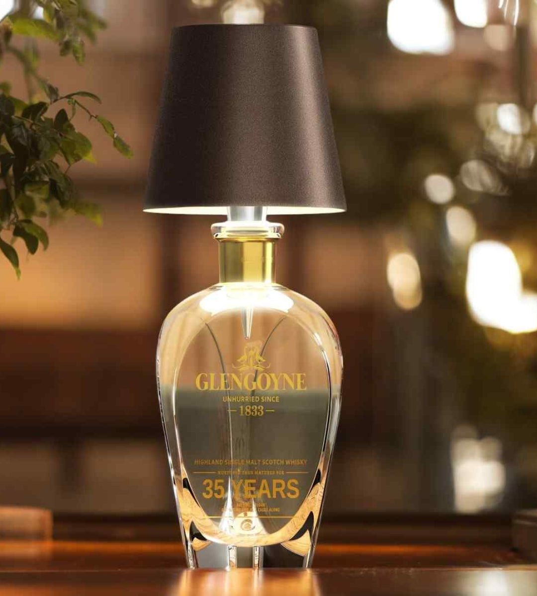 Lampa med mörkbrun skärm på en dekorativ glaskaraff med guldfärgad hals, i varm och dämpad belysning. 5